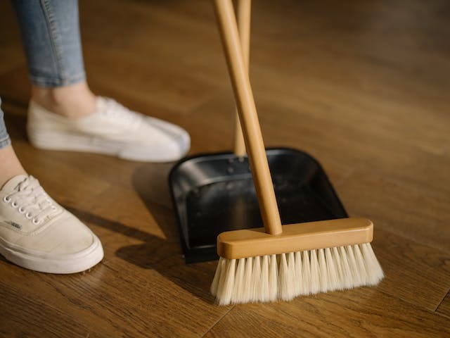 Someone wearing white loafers and jeans (that is all you can see of them) sweeping with a broom and a dustpan on a stick.