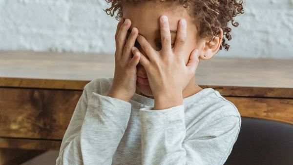 A young boy with dark skin and curly hair covering his face with his hands.