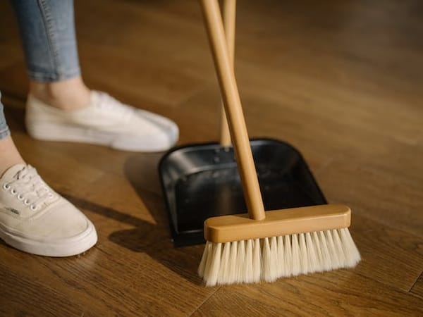 Someone wearing white loafers and jeans (that is all you can see of them) sweeping with a broom and a dustpan on a stick.