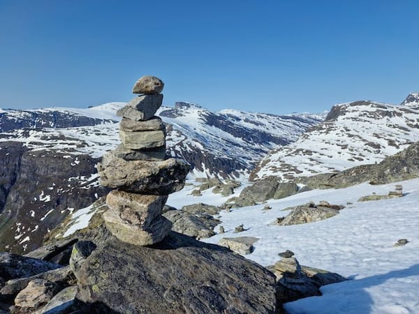 A cairn (stack of rocks) on a mountainside. You can see a valley and some partiall snow covered mountains in the background.